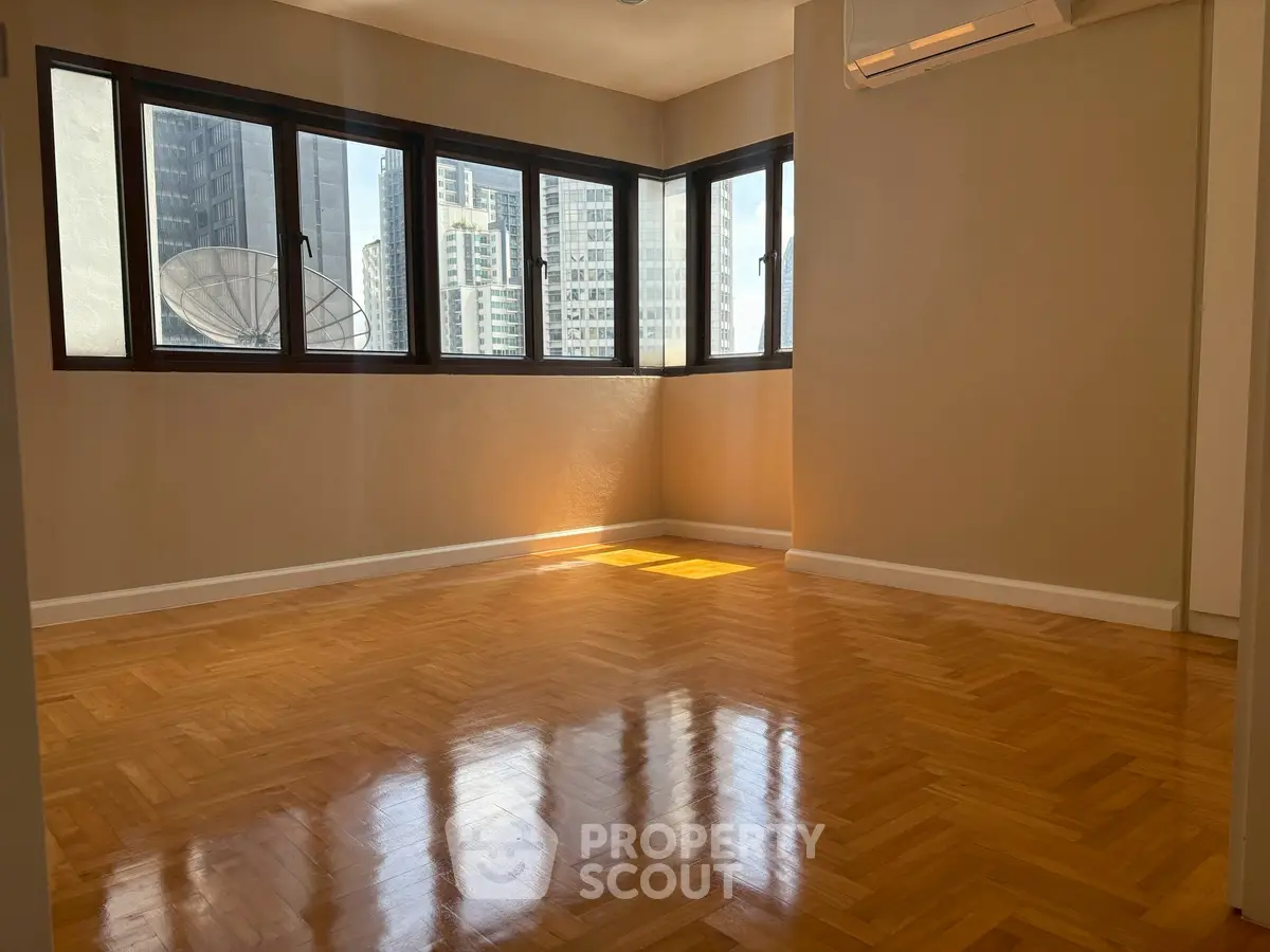 Spacious sunlit room with polished wooden floors and large windows offering city views.