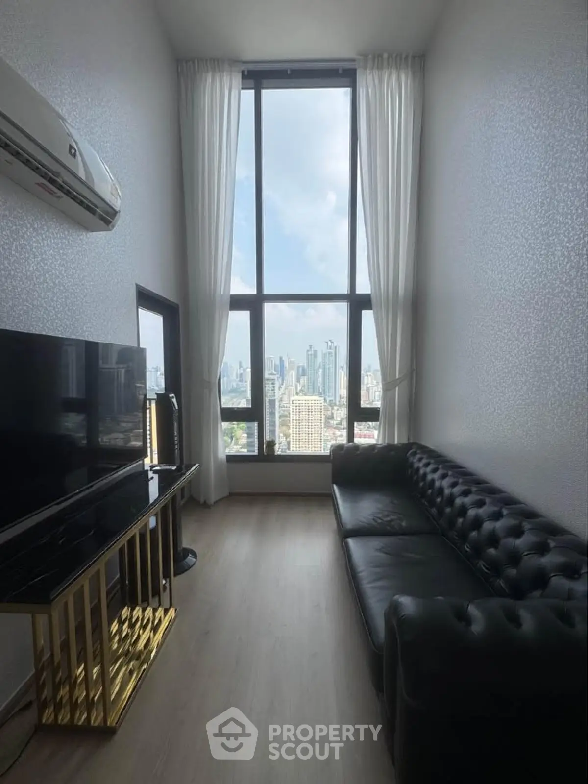Modern living room with high ceiling and city view, featuring sleek black leather sofa and large window.