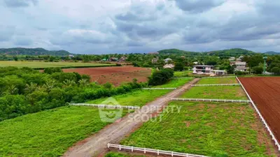 Scenic aerial view of lush green farmland with distant houses and hills under cloudy sky.