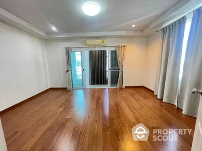 Spacious and well-lit living room with polished wooden flooring, large windows draped with elegant curtains, and a glass door leading to a balcony, perfect for modern living.