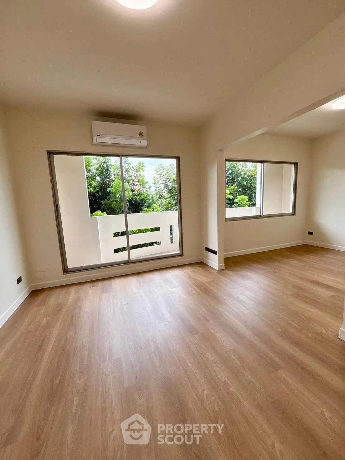 Spacious living room with wooden flooring and large windows offering lush greenery views.