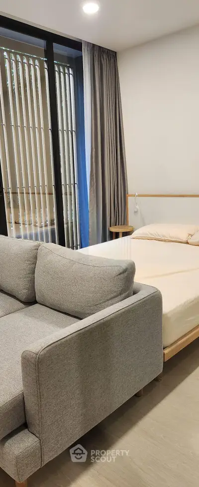 Modern studio apartment with cozy bed and stylish gray sofa near large window with blinds.