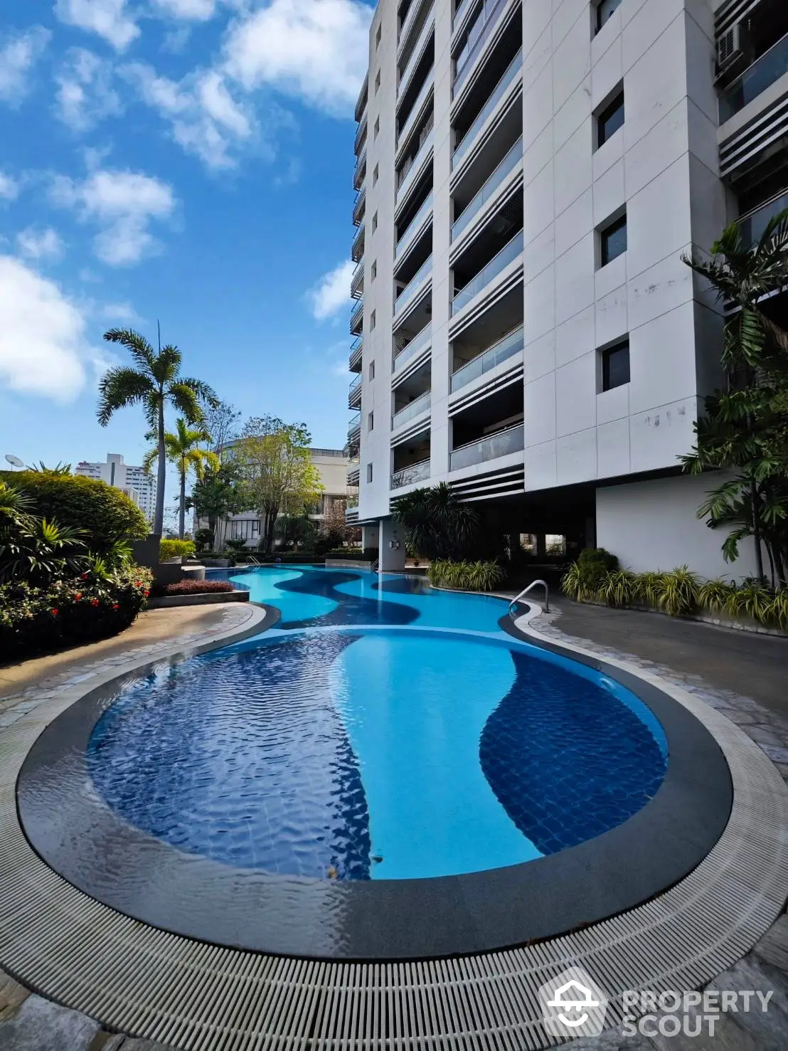 Luxurious residential complex with a stunning blue swimming pool surrounded by lush tropical landscaping and modern high-rise buildings.