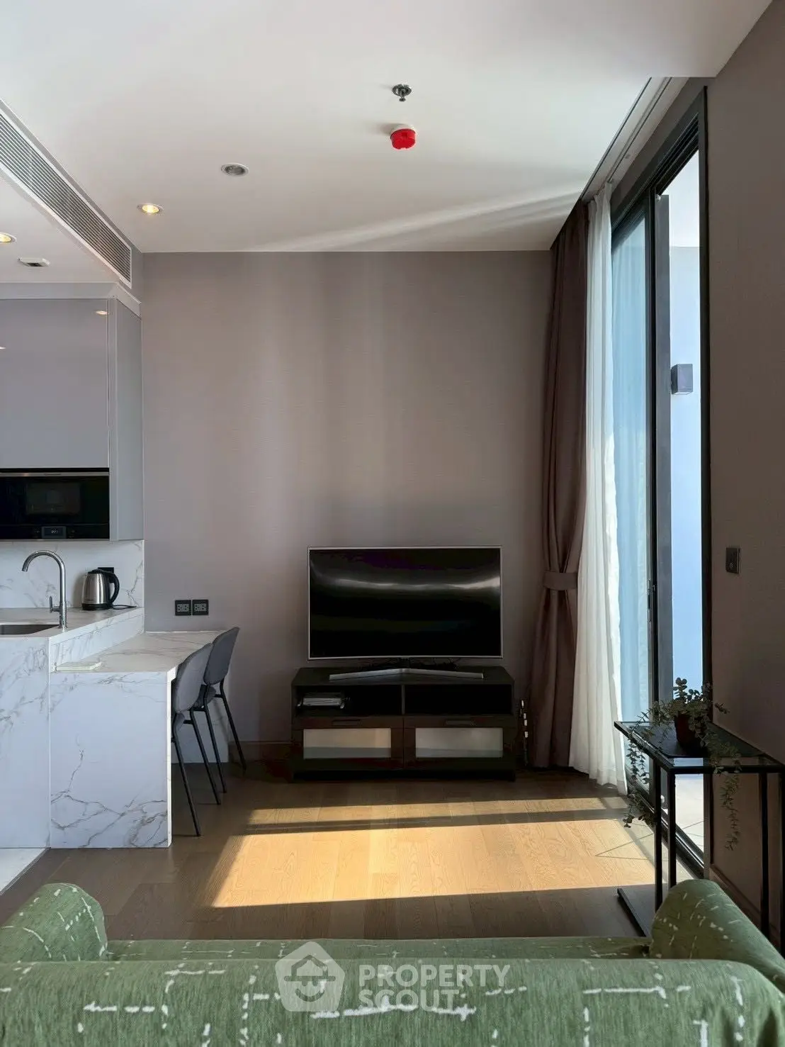 Modern living room with sleek TV setup and natural light, featuring a stylish kitchen counter and floor-to-ceiling windows.