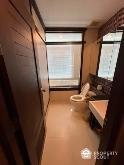 Modern bathroom with sleek fixtures, including a bathtub, toilet, and sink, complemented by warm lighting and a large window with blinds.