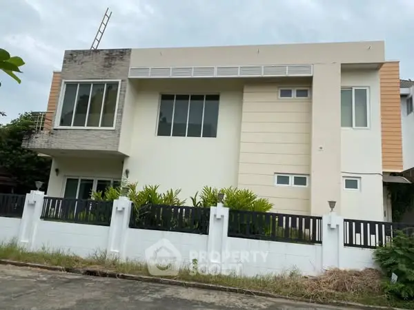 Modern two-story house with large windows and a sleek design, surrounded by a white fence.
