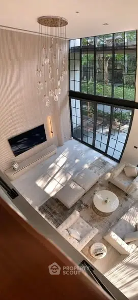Luxurious modern living room with high ceilings, large windows, and elegant chandelier overlooking a serene garden.