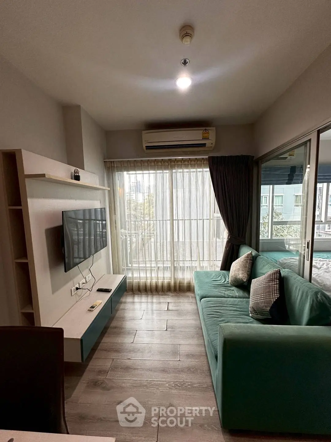 Modern living room with teal sofa, wall-mounted TV, and large window with sheer curtains.