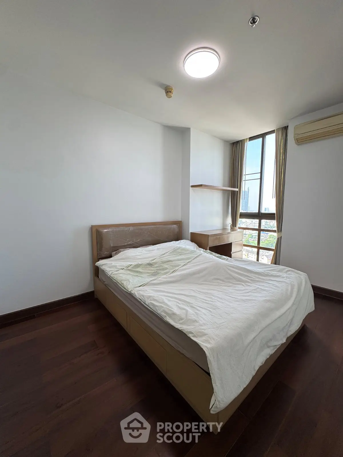 Spacious bedroom with large window offering city views, featuring a comfortable bed and modern wooden flooring.
