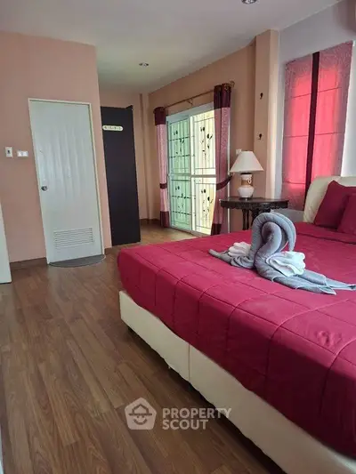 Cozy bedroom with elegant red bedding and wooden flooring, featuring natural light from large windows.
