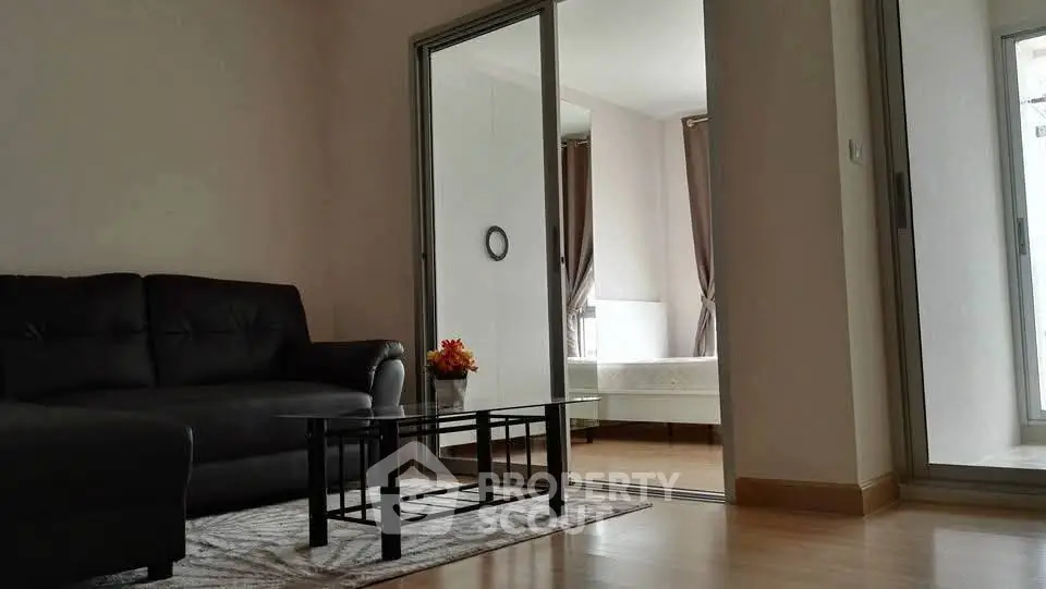 Modern living room with sleek black sofa and glass coffee table, leading to a cozy bedroom.