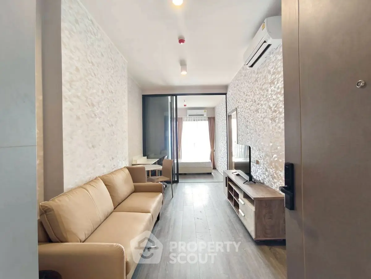 Modern living room with beige sofa, wooden flooring, and air conditioning, leading to a bright bedroom.