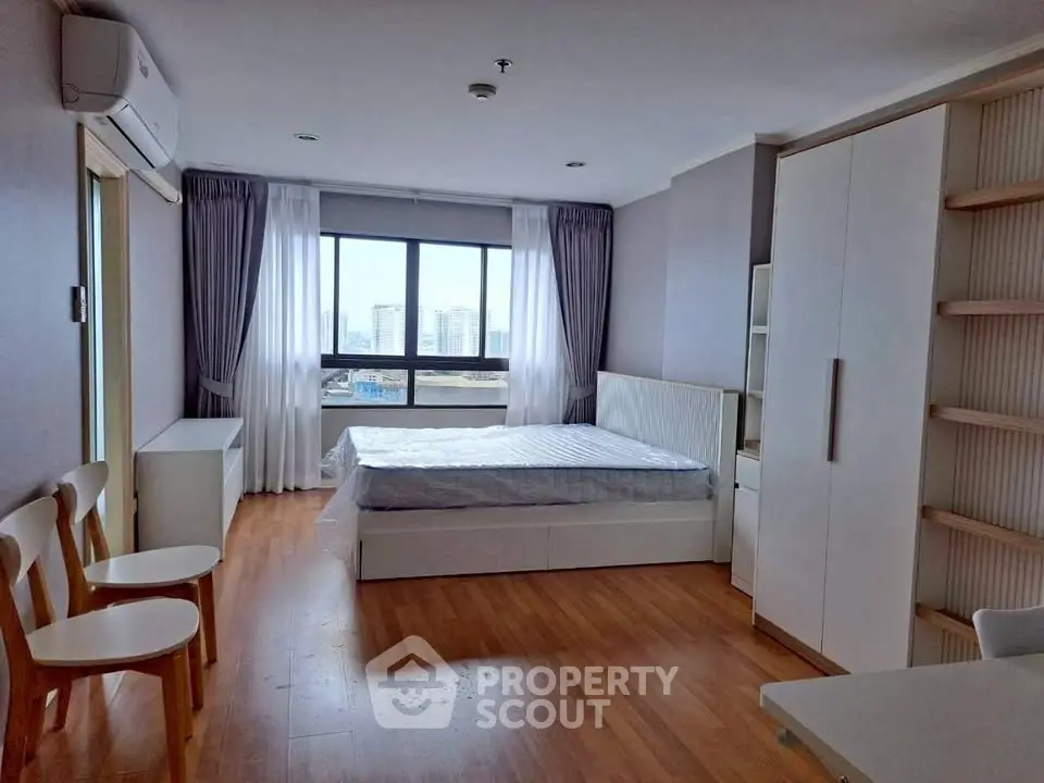 Spacious bedroom with large window offering city views, featuring modern furniture and wooden flooring.