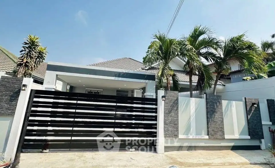 Modern tropical house with sleek gate and lush palm trees under clear blue sky.