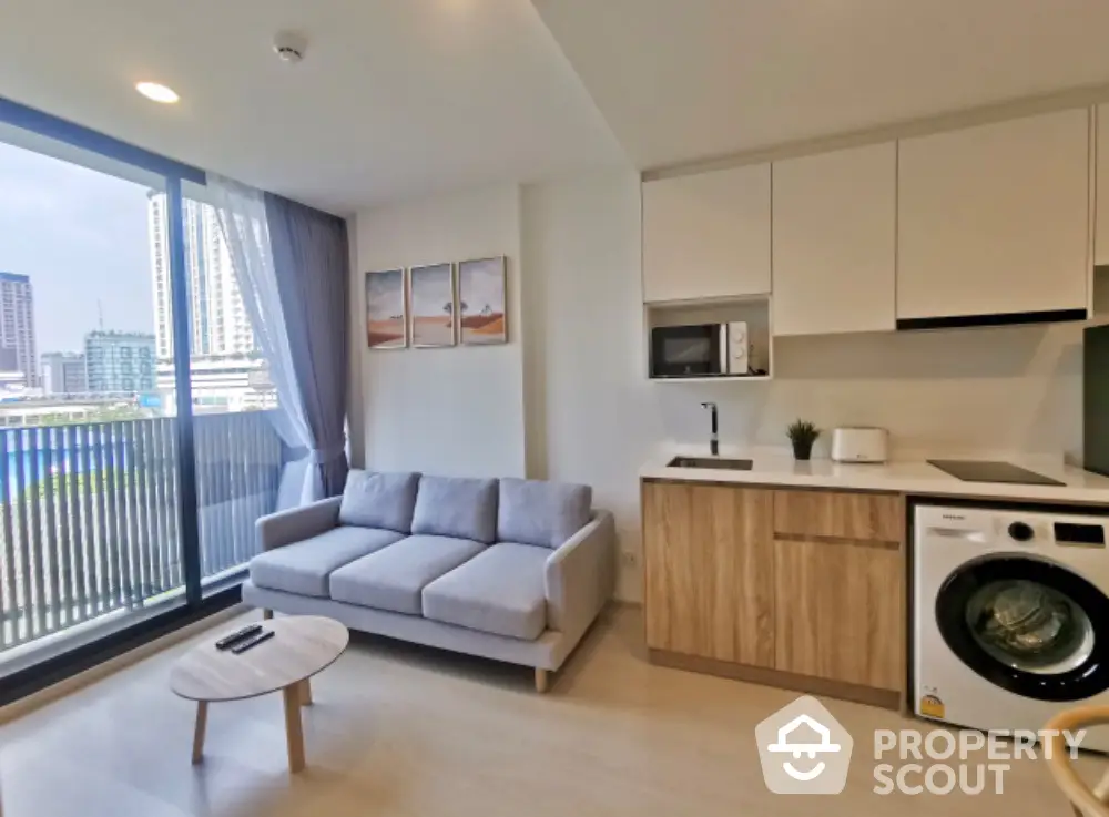 Modern studio apartment with open layout kitchen, cozy living area, and large windows leading to a sunny balcony with urban views.