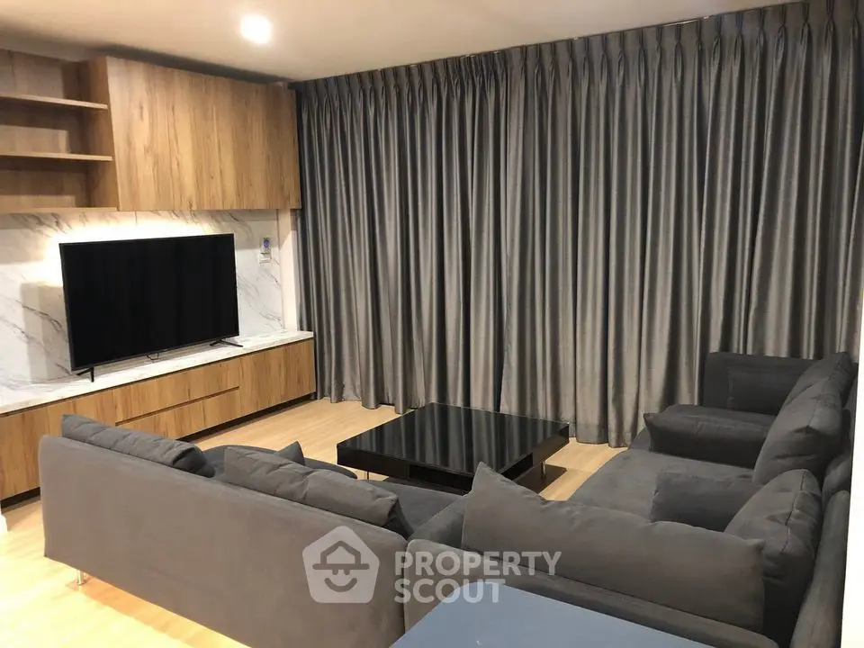 Modern living room with sleek gray sofa and large TV, featuring elegant wooden accents and floor-to-ceiling curtains.