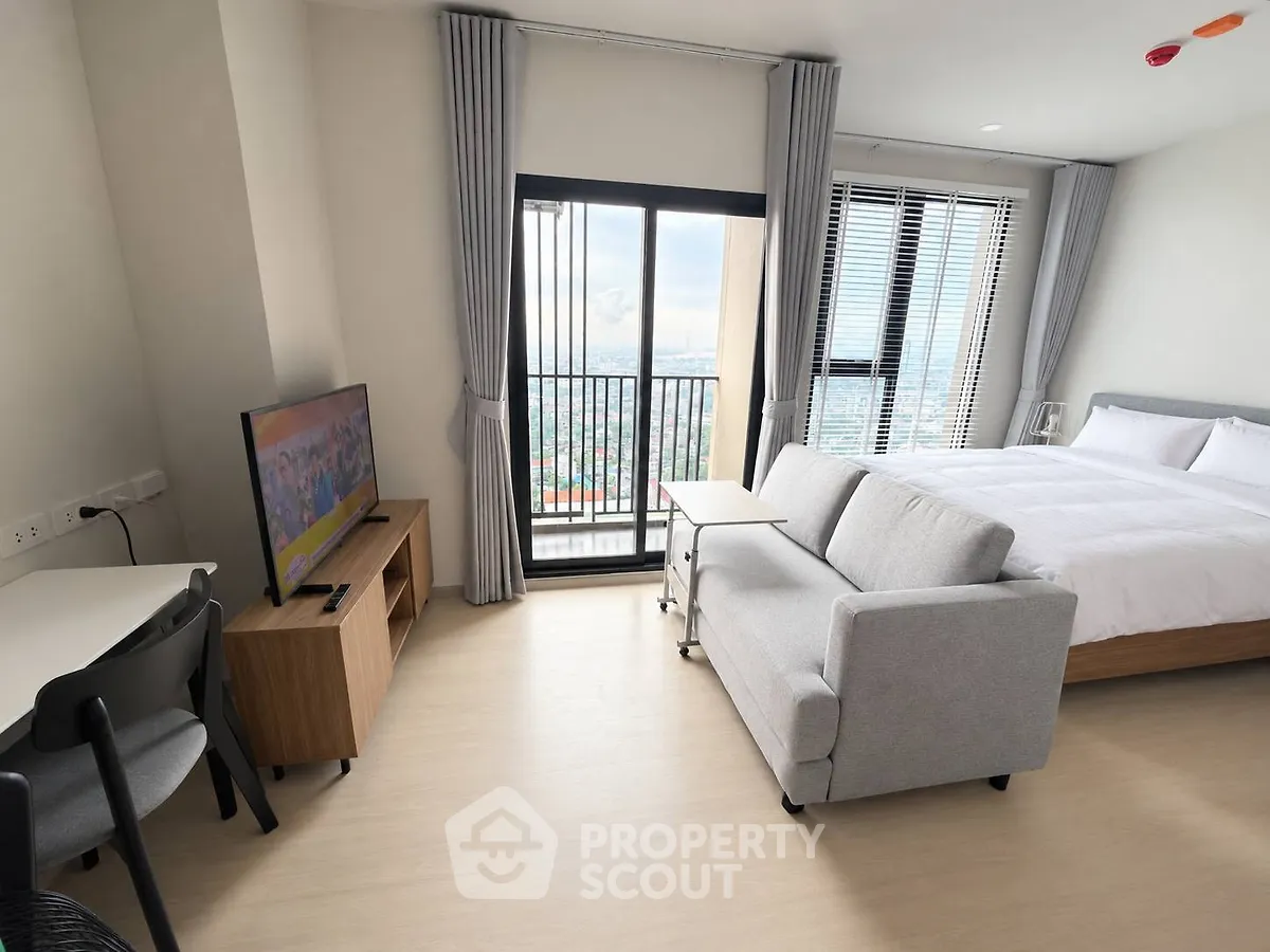Modern studio apartment with balcony and city view, featuring cozy bed, sofa, and TV setup.