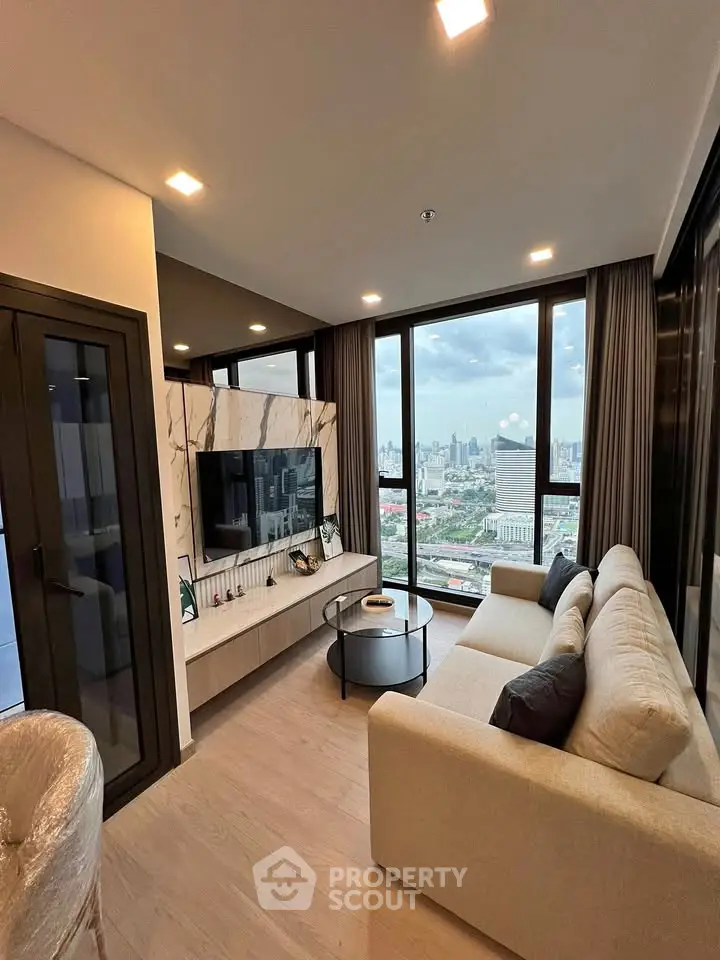 Modern living room with large windows offering stunning city views, featuring a cozy sofa and sleek TV setup.