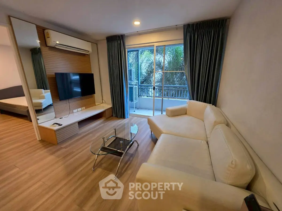 Modern living room with beige sofa and glass coffee table, featuring a wall-mounted TV and balcony view.