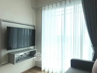 Modern living room with wall-mounted TV and large window with sheer curtains.