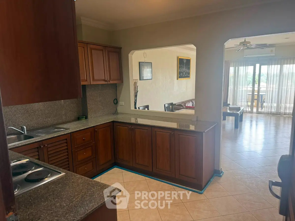 Spacious open-layout kitchen with wooden cabinets and granite countertops leading to a bright living area with large windows.