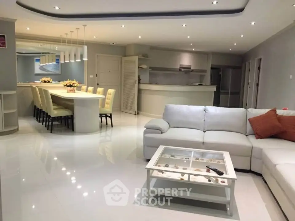 Spacious modern living room with open kitchen and elegant dining area, featuring sleek white furnishings and ambient lighting.