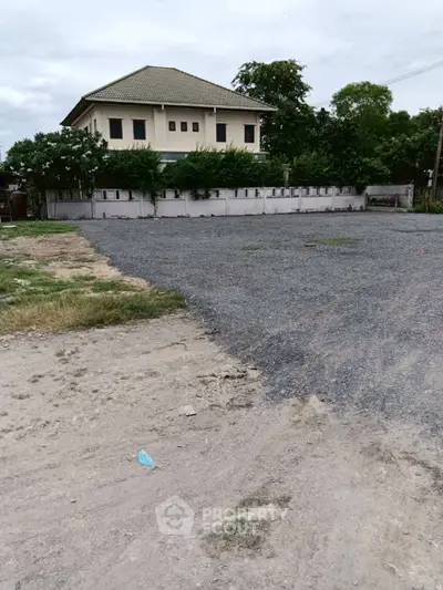 Spacious plot of land with gravel surface and a two-story house in the background, ideal for development.