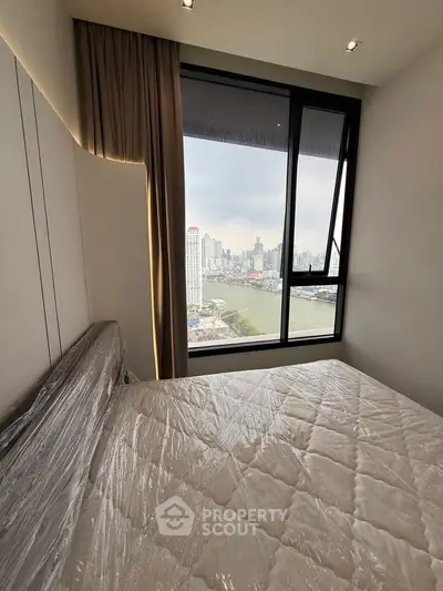Modern bedroom with city view and large window, featuring a wrapped mattress.