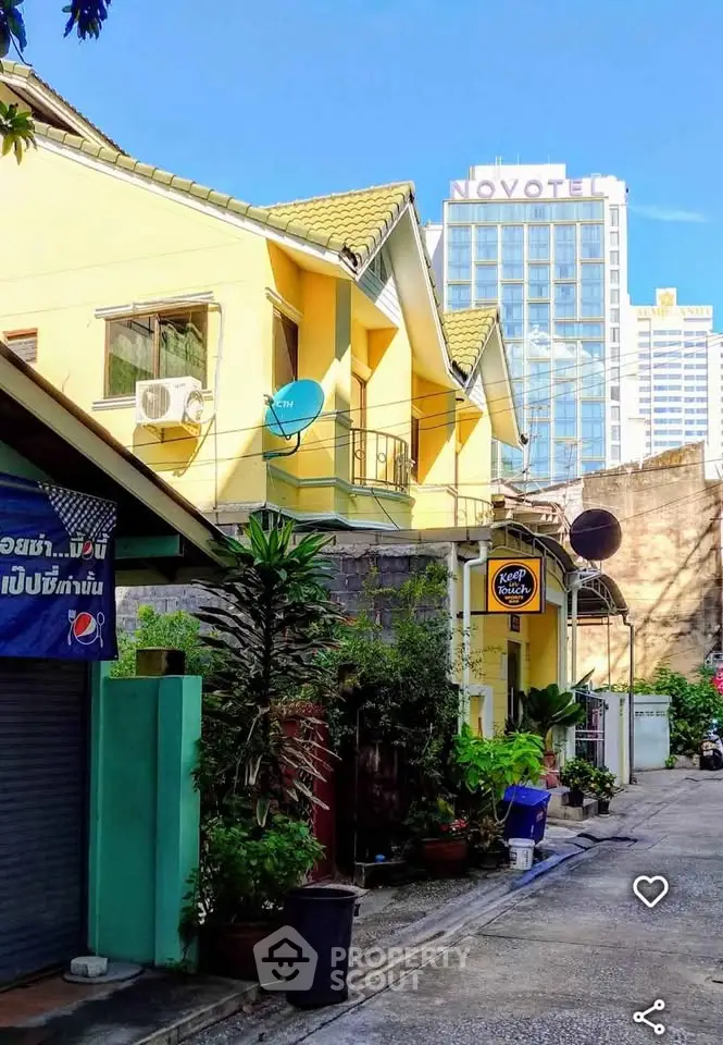 Charming yellow house with lush greenery in urban setting, near high-rise buildings.