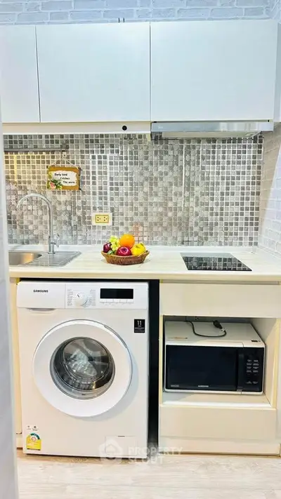 Modern compact kitchen with washing machine and microwave, stylish backsplash, and fruit basket.