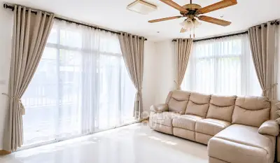 Spacious living room with large windows and elegant curtains, featuring a comfortable beige sofa and ceiling fan.