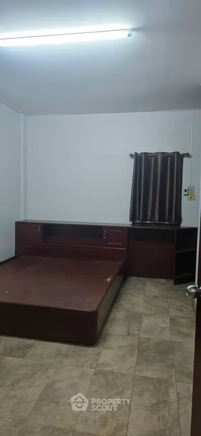 Spacious bedroom with minimalist design and tiled flooring, featuring a built-in wooden bed frame and dark curtains.