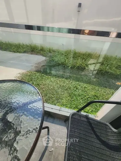 Charming garden view from a cozy balcony with glass table and chair, perfect for relaxation.