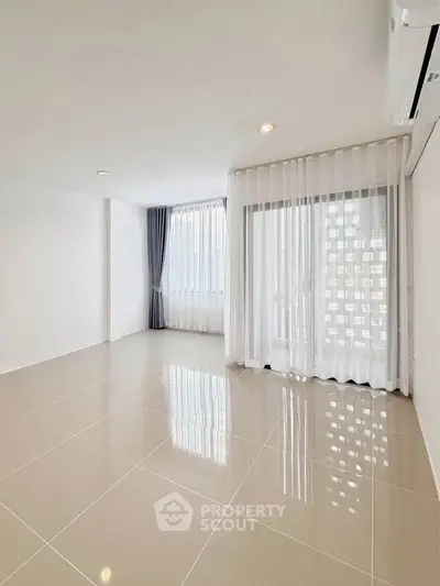 Spacious empty room with glossy tiled floor and large windows with sheer curtains.