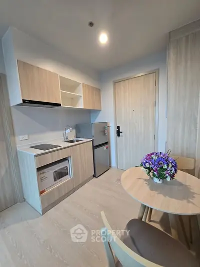 Modern compact kitchen with light wood cabinets and dining area, featuring a small fridge and microwave.