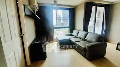 Modern living room with large windows and city view, featuring a sleek gray sofa and wooden flooring.