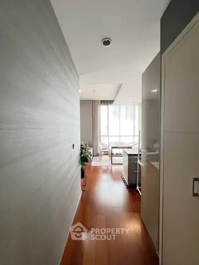 Modern apartment hallway leading to a bright living area with wooden flooring and large windows.