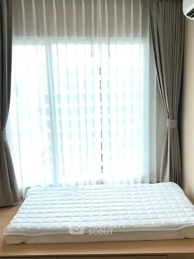 Bright bedroom with large window and elegant curtains, featuring a cozy mattress on a wooden platform.