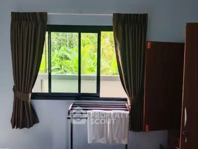 Cozy bedroom with window view of lush greenery, featuring dark curtains and a towel rack.