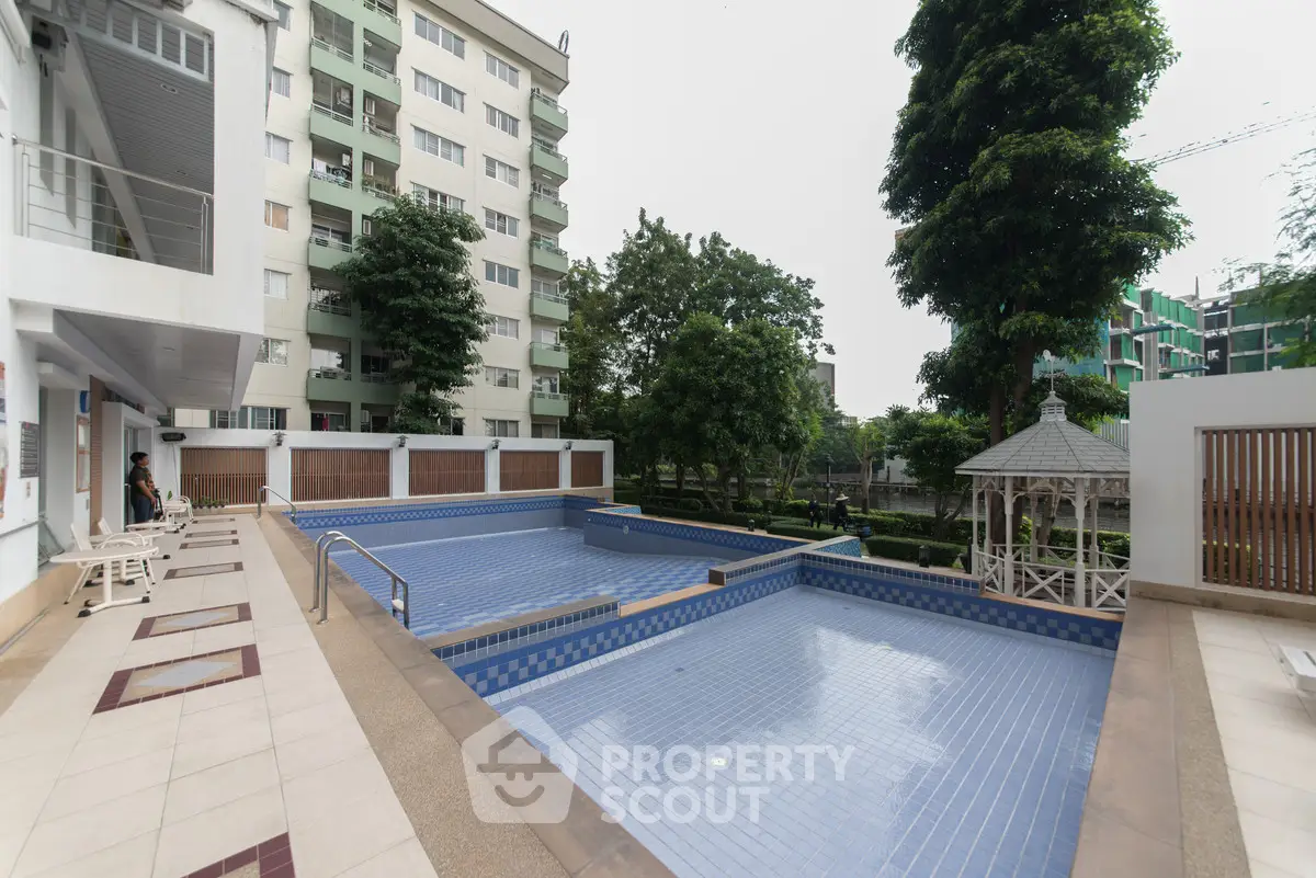 Spacious outdoor pool area with modern design in a residential complex, surrounded by lush greenery and elegant architecture.
