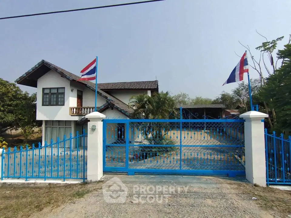 Charming two-story house with blue gate and flags, spacious driveway, lush greenery, and serene ambiance.