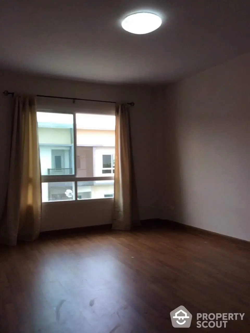 Spacious and well-lit empty room with hardwood flooring and large window draped with golden curtains, ready for personalization.