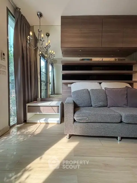 Modern living room with elegant chandelier and cozy gray sofa, featuring wooden flooring and large windows.