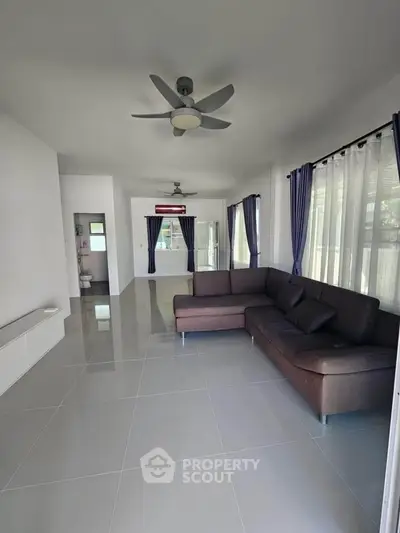 Spacious living room with modern sofa and ceiling fans, featuring large windows and elegant curtains.