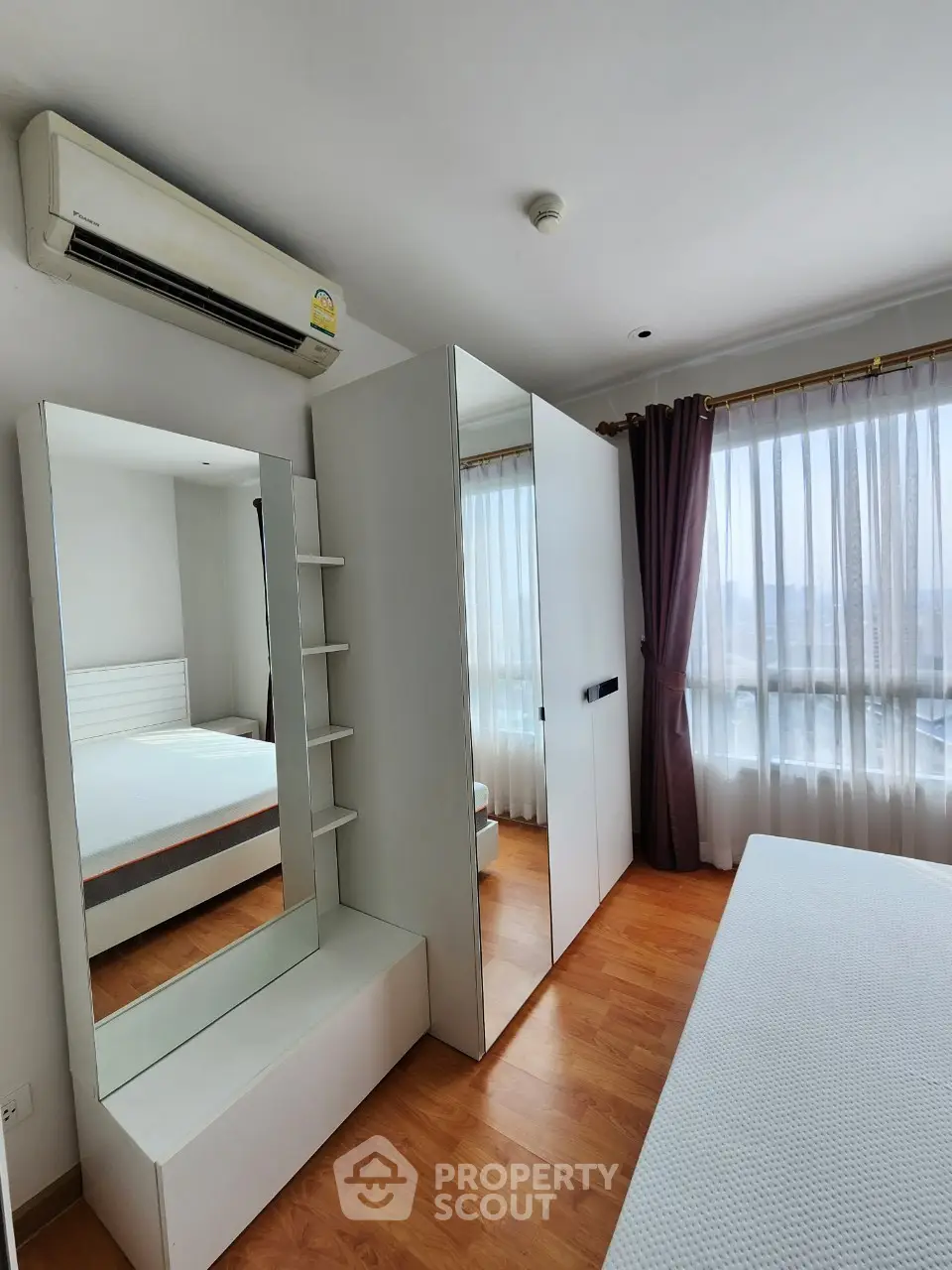 Bright bedroom with large mirror and wardrobe, featuring wooden flooring and air conditioning.