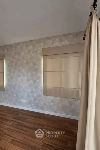 Spacious room with elegant wallpaper and wooden flooring, featuring large windows with beige curtains.