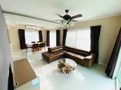 Spacious living room with elegant leather sofa and dining area, featuring large windows and modern ceiling fan.