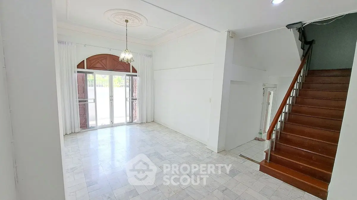 Spacious living room with elegant chandelier and marble flooring, featuring a wooden staircase and large windows.