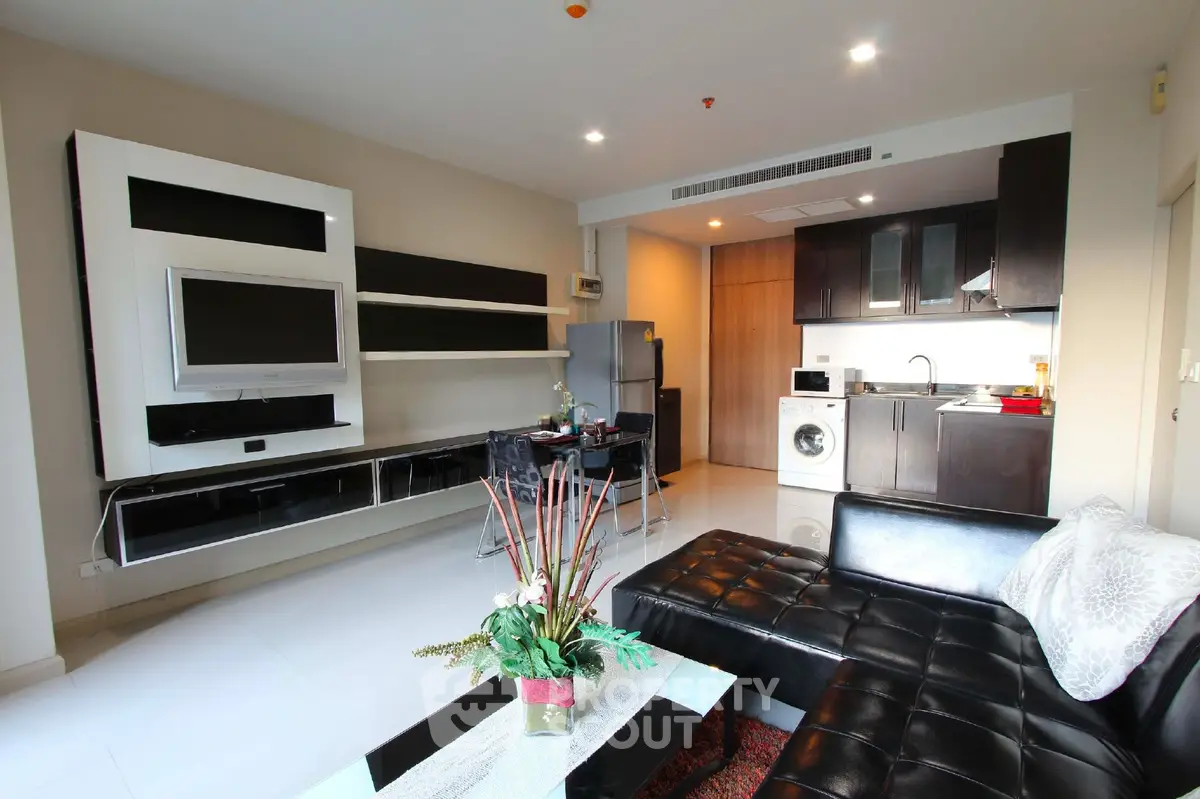 Modern living room with sleek black furniture and open kitchen layout, featuring a washing machine and flat-screen TV.