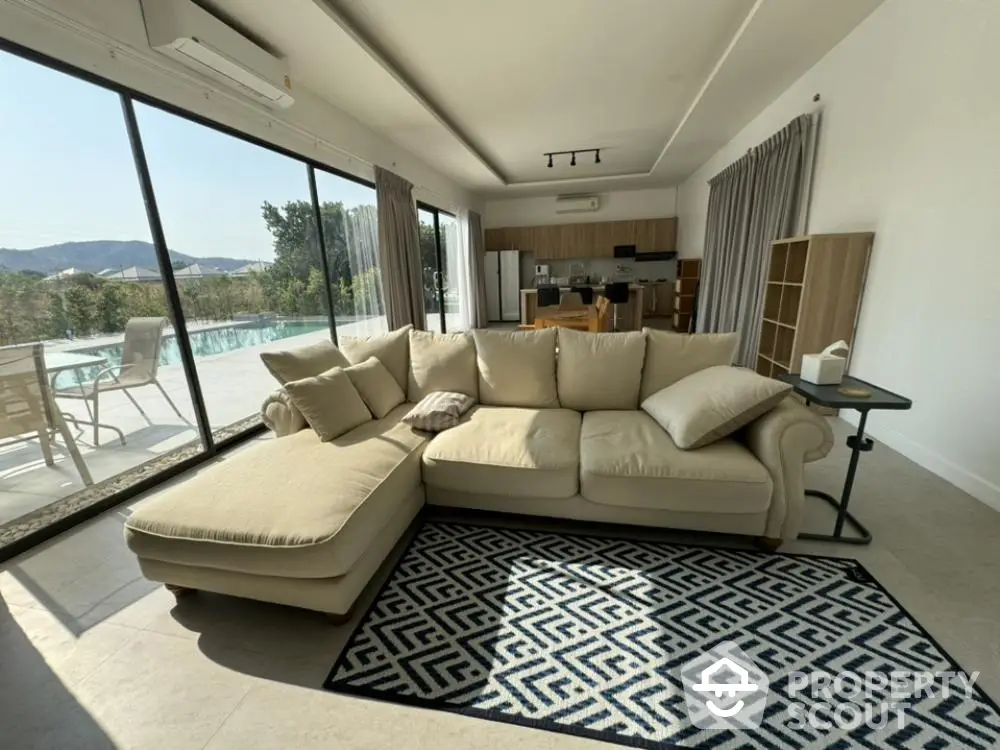 Spacious living room with large sectional sofa and geometric rug, opening to a sunlit balcony with a serene pool view, perfect for luxurious relaxation.